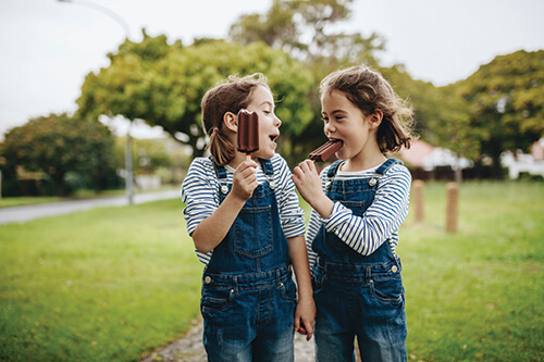 Two little twin sisters in identical clothes standing outdoors and eating chocolate ice cream candy.